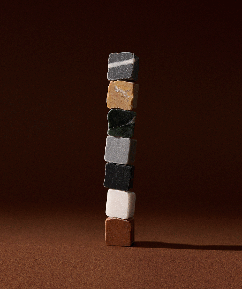 A vertical stack of seven small stone or marble cubes in various colors, including gray, beige, green, black, white, and brown, against a brown background.