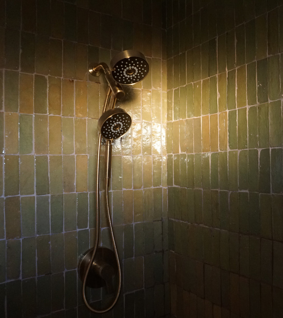 A wall-mounted shower fixture with a handheld showerhead against a wall of glossy, rectangular green Za’atar 2x6 tiles.