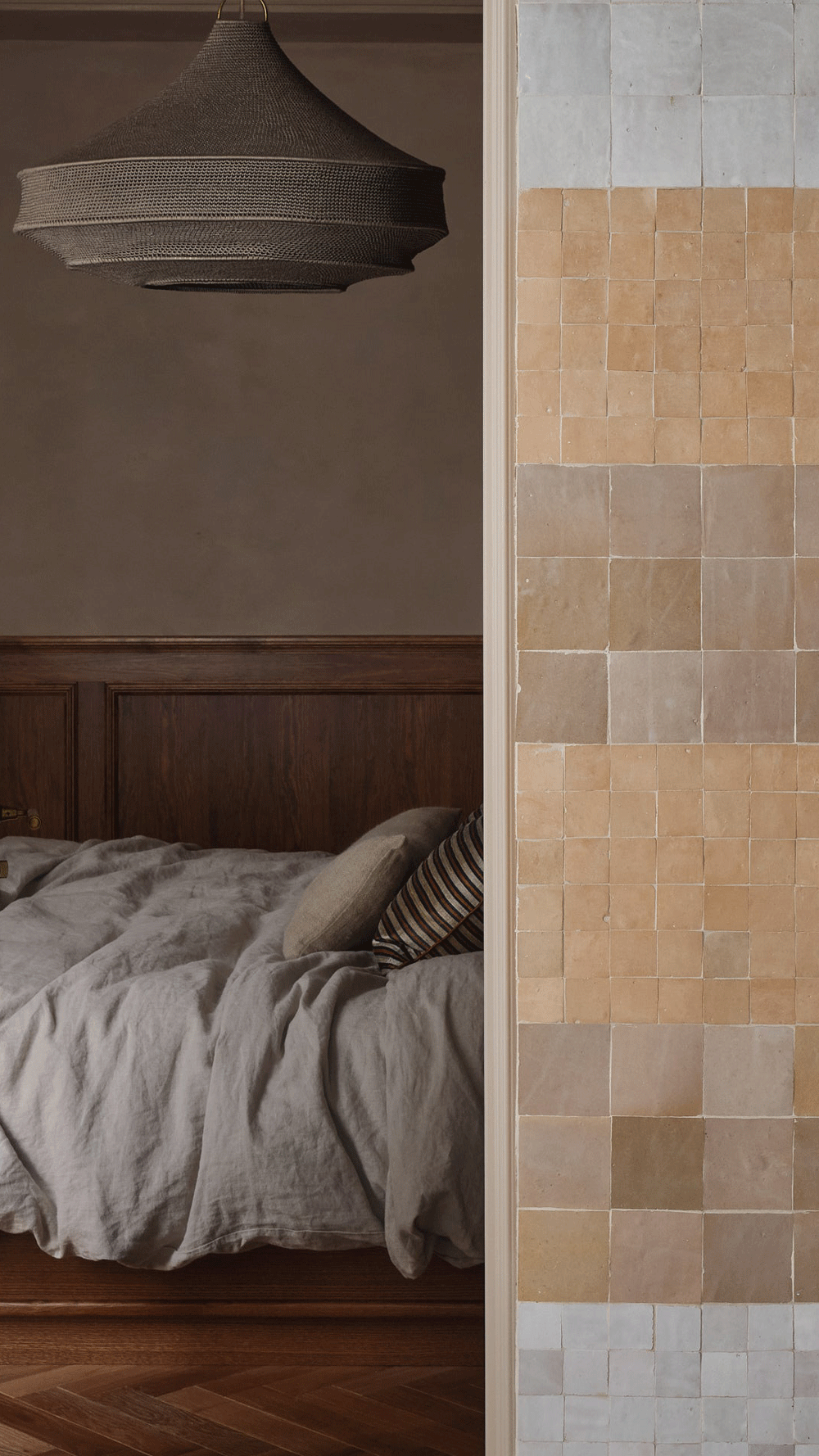 A cozy bedroom with a linen-covered bed, wooden headboard, and neutral-toned pillows, partially visible behind a wall tiled with beige and light gray squares; a woven pendant light hangs from the ceiling.
