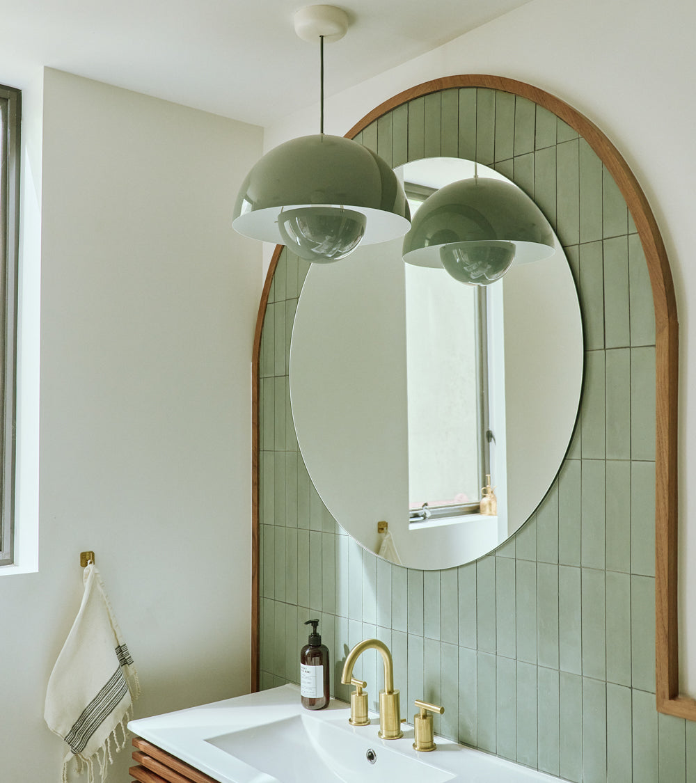 Modern bathroom featuring vendor-unknown's Zeppelin 2x8 green subway tile, round mirror, gold faucet, white sink, soap dispenser, wall-mounted towel, and a large dome-shaped pendant light reflected in the mirror.