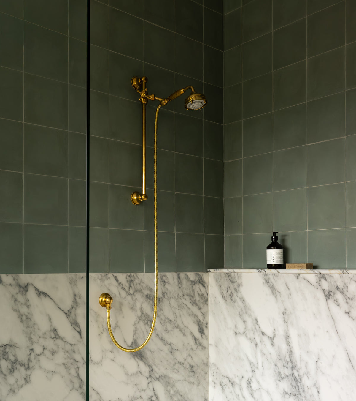 A shower featuring Zeppelin 8x8 green cement tiles (vendor-unknown) on the upper walls, white marble tiles below, and a gold shower fixture. A bottle and a small item rest on a corner shelf.