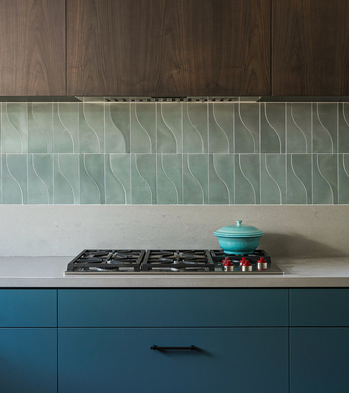A modern kitchen features the Nouveau Zeppelin by vendor-unknown, a gas stovetop with red knobs, blue lower cabinets, dark wood uppers, and a green wavy-pattern cement tile backsplash.