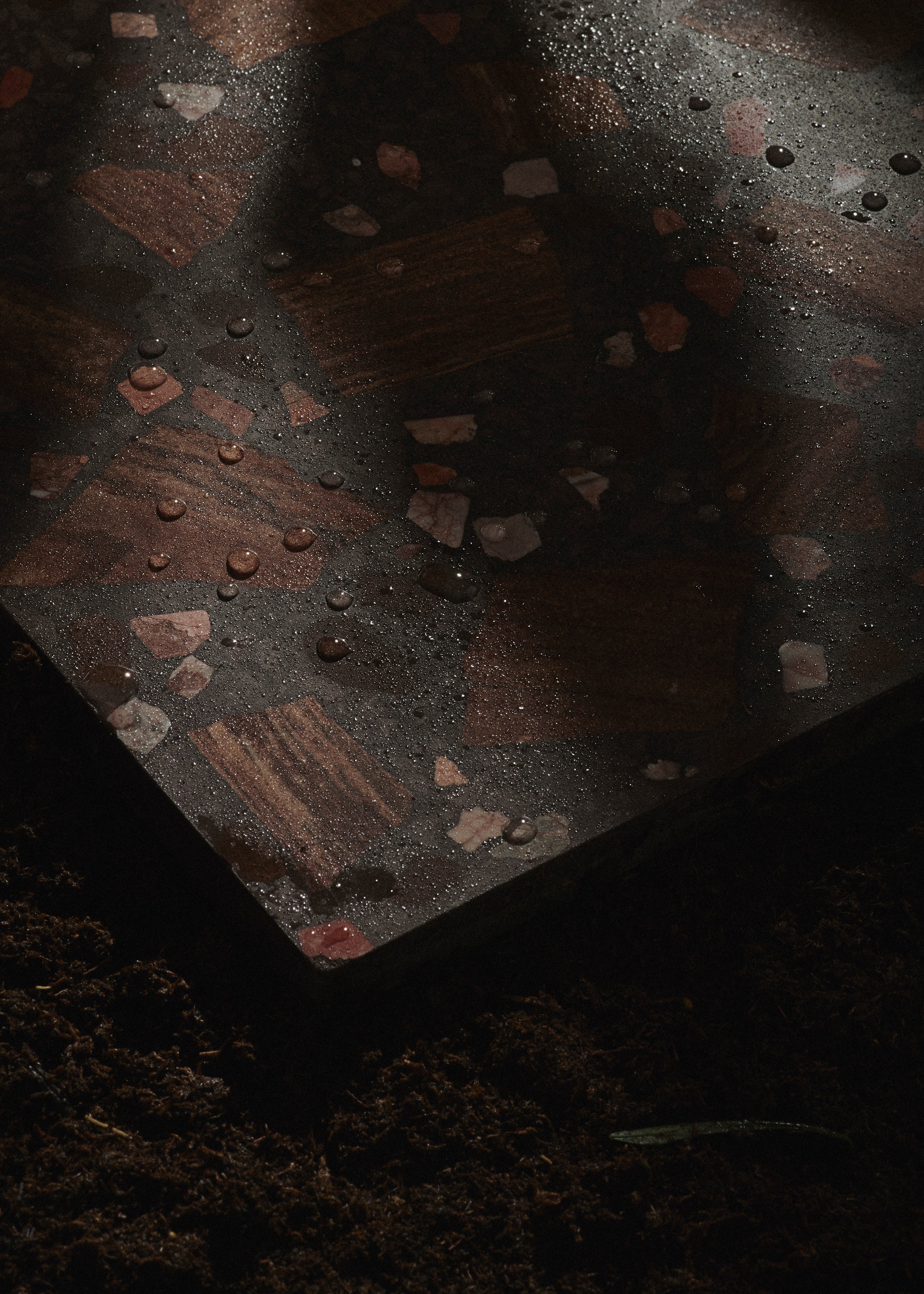 A dark, patterned stone tile with scattered water droplets rests on soil. The tile features various shades of brown and pink in a mosaic design, and dramatic shadows partially cover its surface.