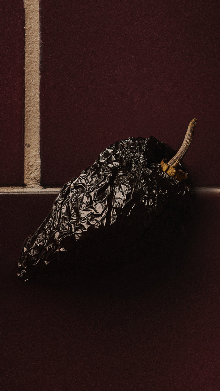 A dried dark chili pepper with a wrinkled surface and a short stem rests on a maroon tiled surface with beige grout lines.