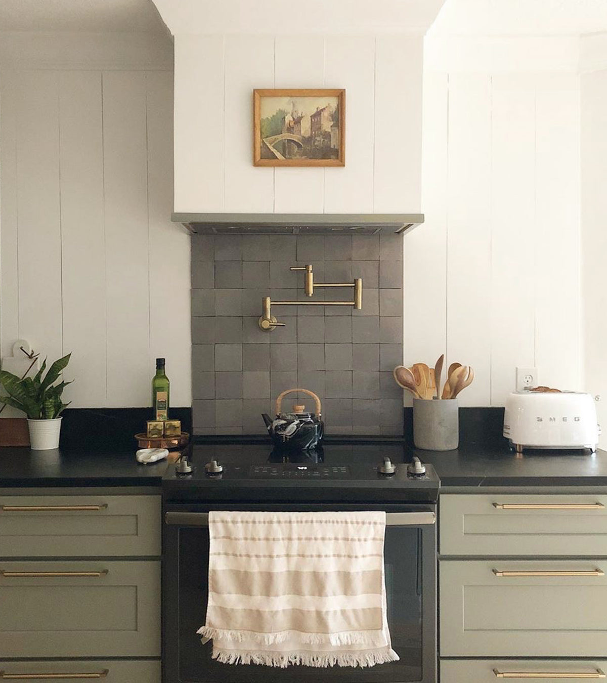 A modern kitchen with sage green cabinets, black countertop, and a Zinc 4x4 gray Moroccan tile backsplash by vendor-unknown, featuring a stove with striped towel, kettle, utensils, toaster, plant, olive oil, and framed painting on a white wall.