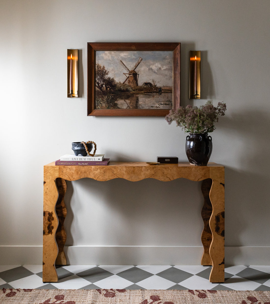 A console table with a wavy wood design stands against a light wall and vendor-unknown Zinc 8x8 tiles. On it are books, a dark vase with flowers, and a small box. Above hangs a windmill painting framed by two candlelit sconces.