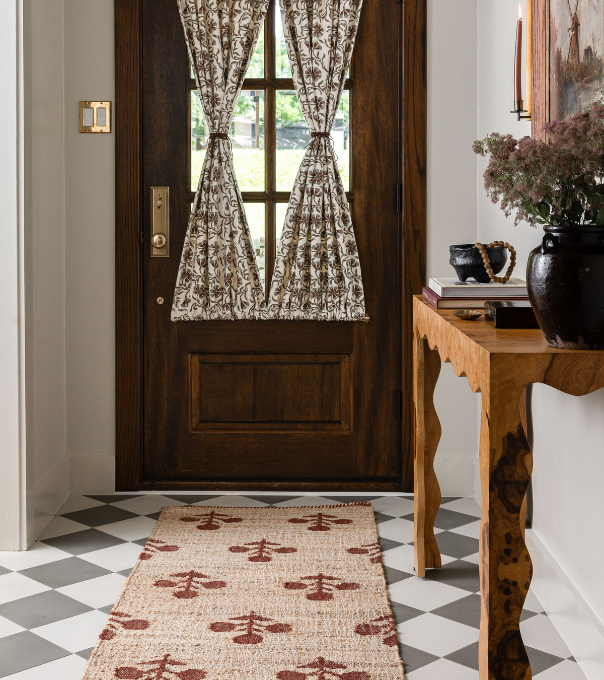 A wooden front door with patterned curtains is shown in a bright entryway featuring Zinc 8x8 cement tile by vendor-unknown in a black and white checkered pattern. A patterned rug leads to a wood console table with vases, books, and flowers.