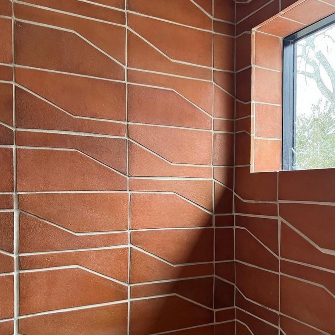 A corner of a room showcases Zia Tile’s Zocalo + Red Clay terra cotta tiles with white geometric grout lines, placed beside a window that reveals lush greenery outside.