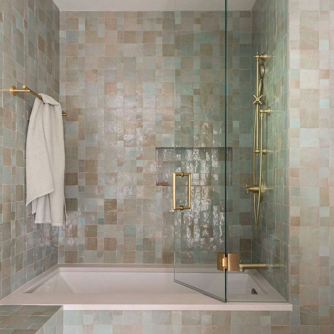 A bathroom with a light green and beige tiled wall, a white bathtub, gold shower fixtures, a glass shower door, and a white towel hanging on a gold towel bar.