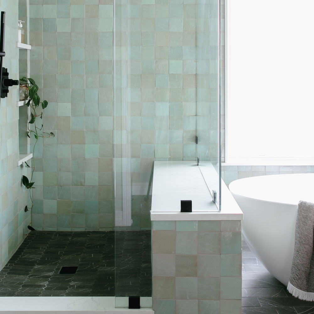 Modern bathroom with greenish square tiles, a glass shower enclosure, a white bathtub, and a towel draped over the tub’s edge. There’s a small plant on a shelf in the shower area. Natural light fills the space.
