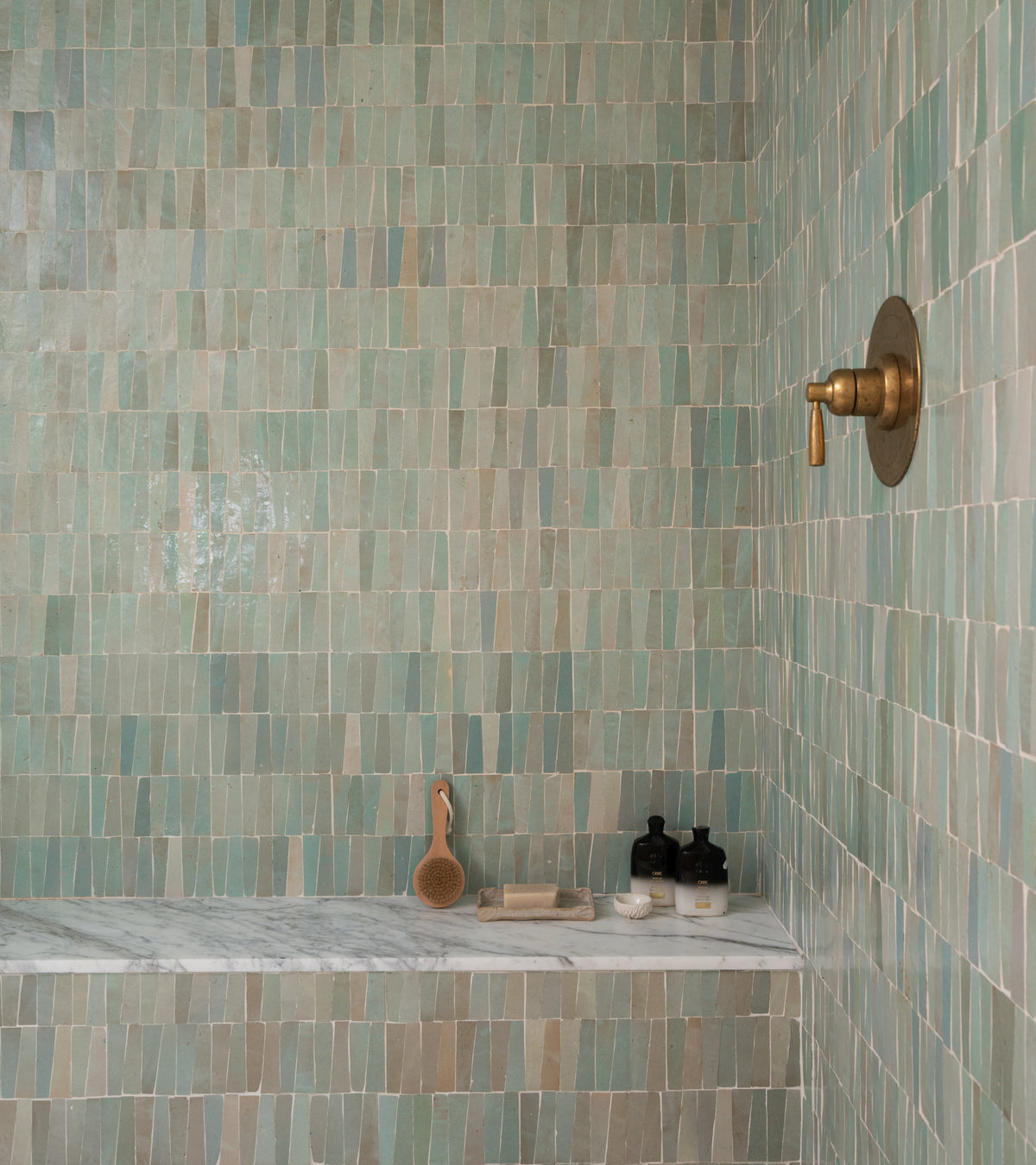 A shower features Zia Tile's Absinthe Trapezoid tiles, a brass showerhead, and a marble bench displaying a wooden brush, two dark bottles, soap, and a dish.