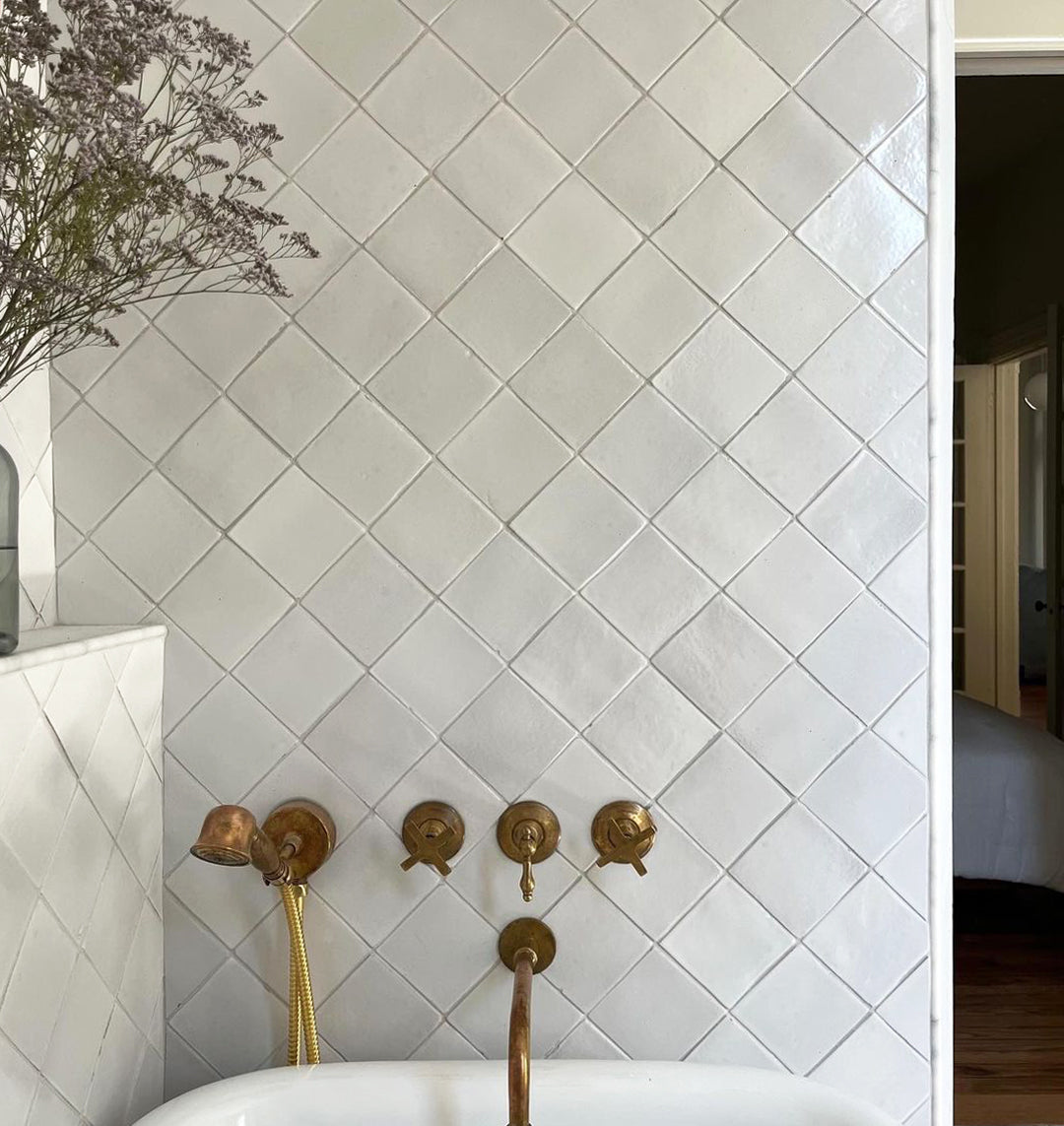 A white bathroom wall showcases Zia Tile’s Albar 4x4 Square tiles, accented with brass fixtures above the tub and a vase of dried flowers; through the doorway, a San Miguel de Allende-inspired bedroom is visible.