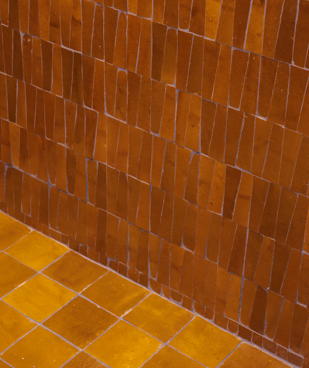 A wall and floor covered with rectangular, reddish-brown tiles arranged in a grid pattern. The tiles on the wall are darker and vary in shade, while the floor tiles are more uniform and yellowish-brown.