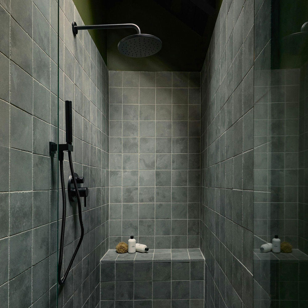 A modern shower with gray square tiles, a black rainfall showerhead and controls, and a built-in tiled bench holding soap, a sponge, and a small bottle.