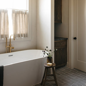 A modern bathroom features a white bathtub, gold faucet, and a small window with beige curtains. A wooden stool with a soap bar and flowers sits beside the tub on Zia Tile’s Basilica 4x12 + Honed limestone flooring.