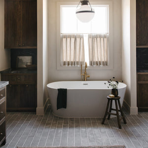 A modern bathroom features a freestanding white bathtub below a window with beige curtains, dark wood cabinets, and Zia Tile's Basilica 4x12 Honed tiles. A gold faucet, dark towel, rug, and floral stool add the finishing touches.