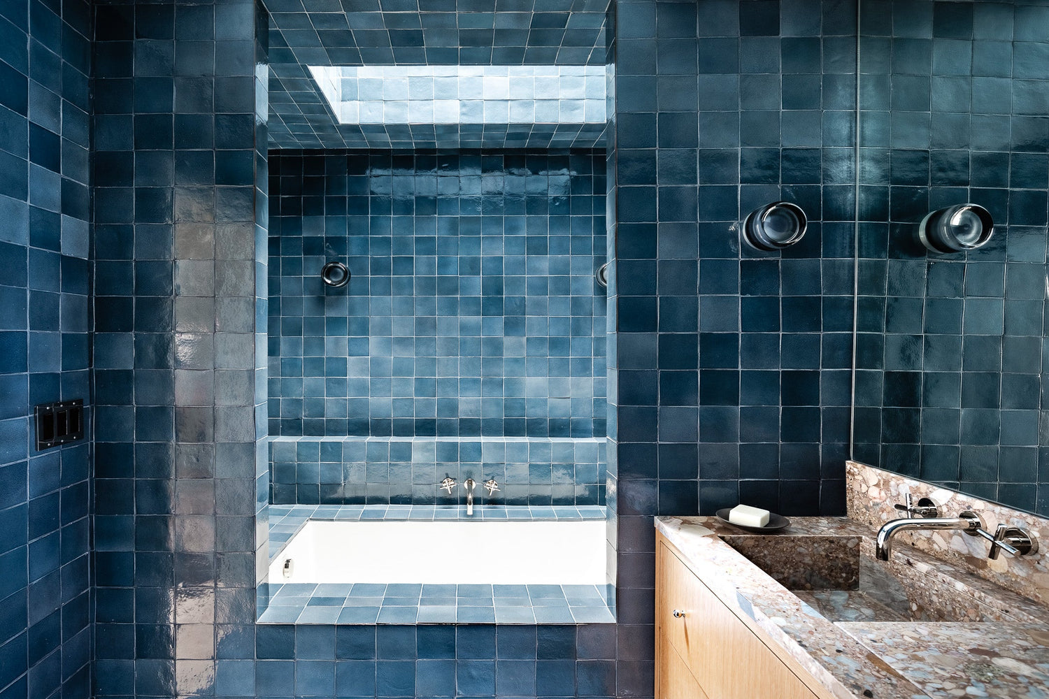 A modern bathroom with glossy blue square tiles covering the walls and ceiling, a white built-in bathtub beneath a skylight, two round wall lights, and a marble countertop with a sink and soap dish.