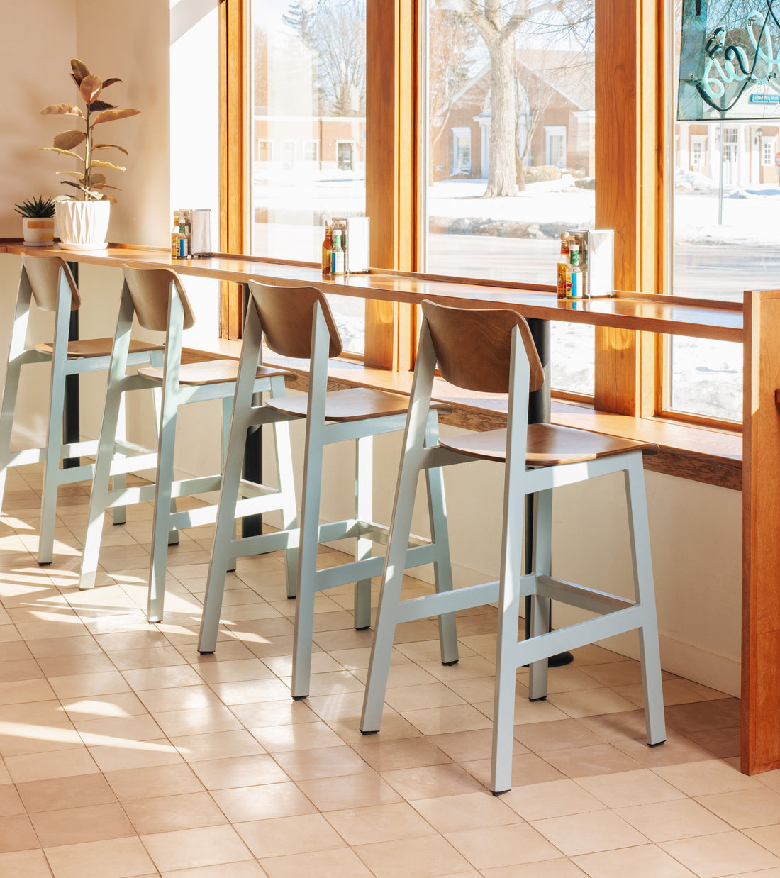 Four light blue and wood bar stools line a high counter by large sunlit windows in a cozy café, where Bone 8x8 tiles from vendor-unknown enhance the charm. Condiment bottles sit on the counter, with a potted plant on the side.