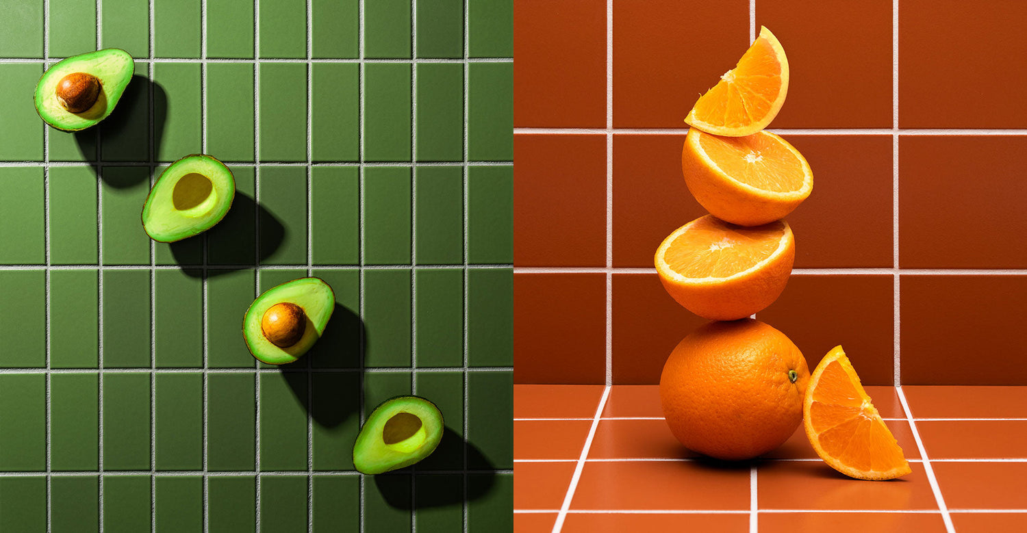 Four halved avocados are arranged diagonally on green tiles, while three stacked oranges and an orange wedge are placed on brown tiles, creating a colorful, geometric composition.