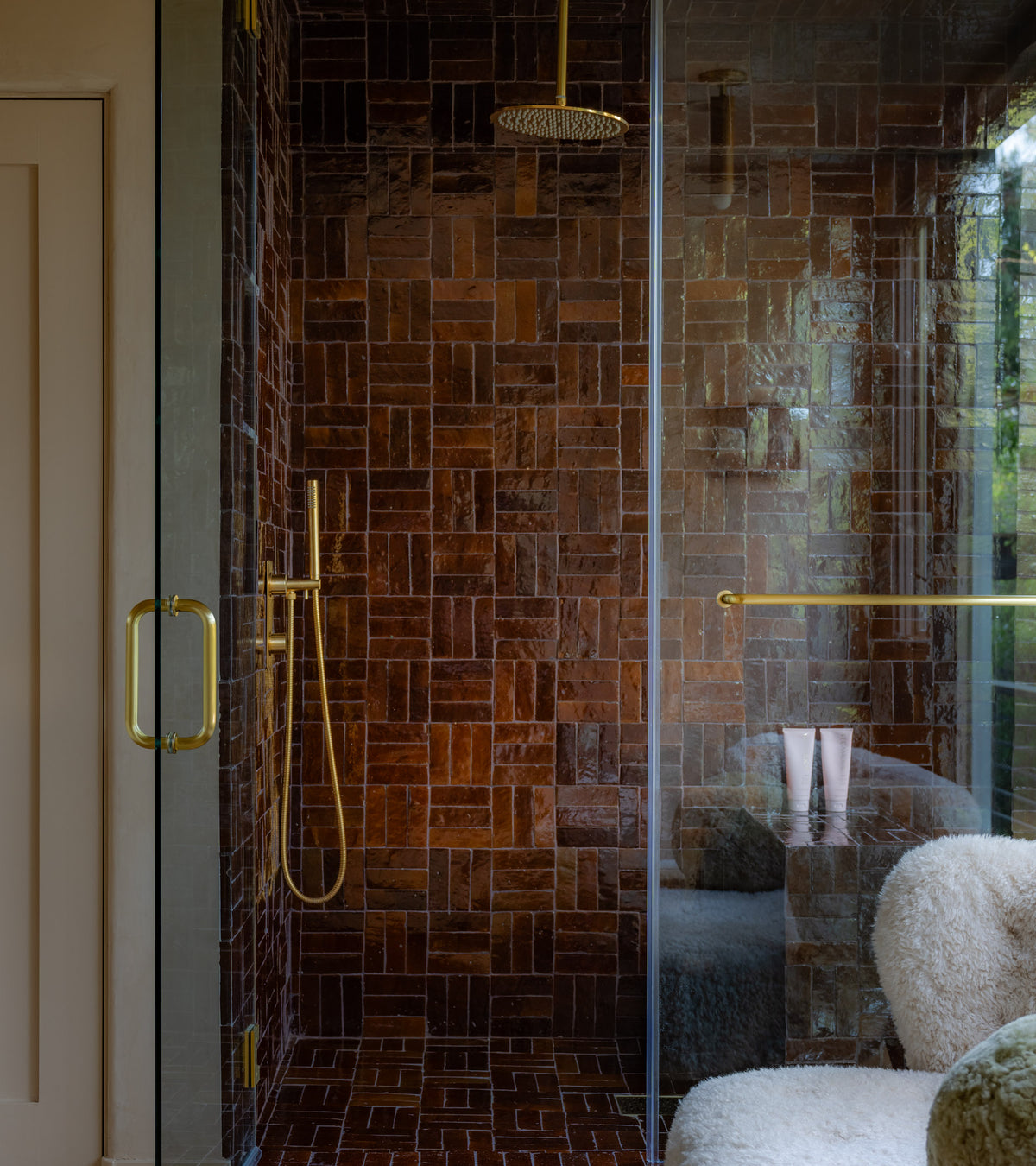 A shower features Zia Tile’s Burnt Sugar 2x6 tiles, a glass door with gold hardware, a rain showerhead, and a handheld. Outside sit a plush white chair and a small table with two pink cups.