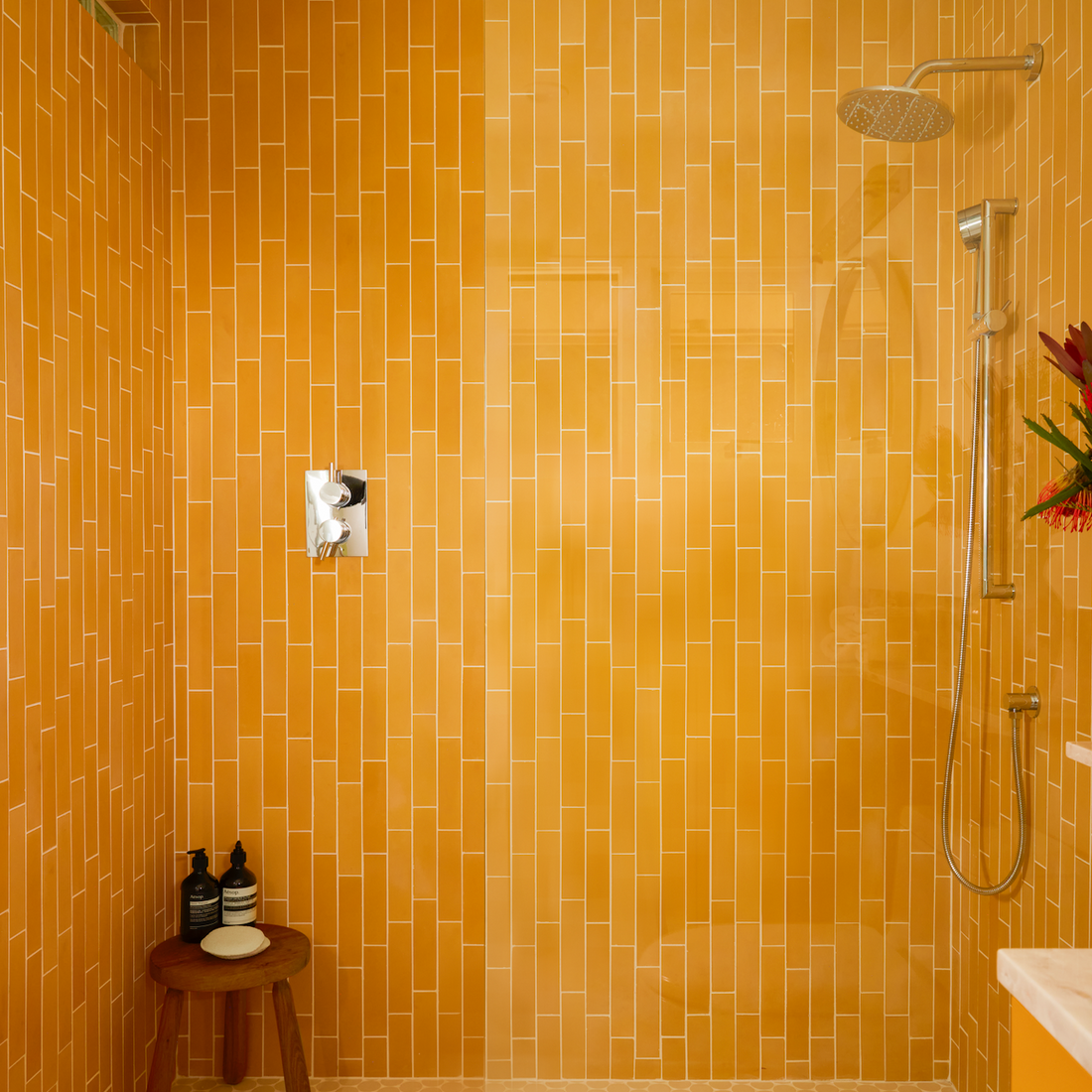 A modern shower area features bright orange vertical subway tiles, a glass partition, a rainfall showerhead, a handheld shower, a wooden stool with toiletries, and hexagonal floor tiles.