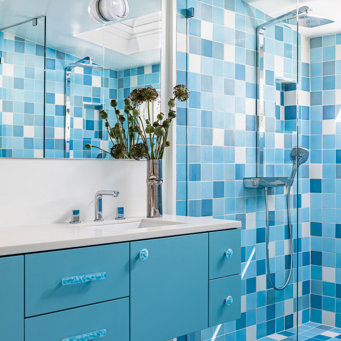 Modern bathroom with blue and white square tiles, floating blue vanity with sink and mirror, a vase of flowers, and a glass-enclosed shower with rainfall showerhead.