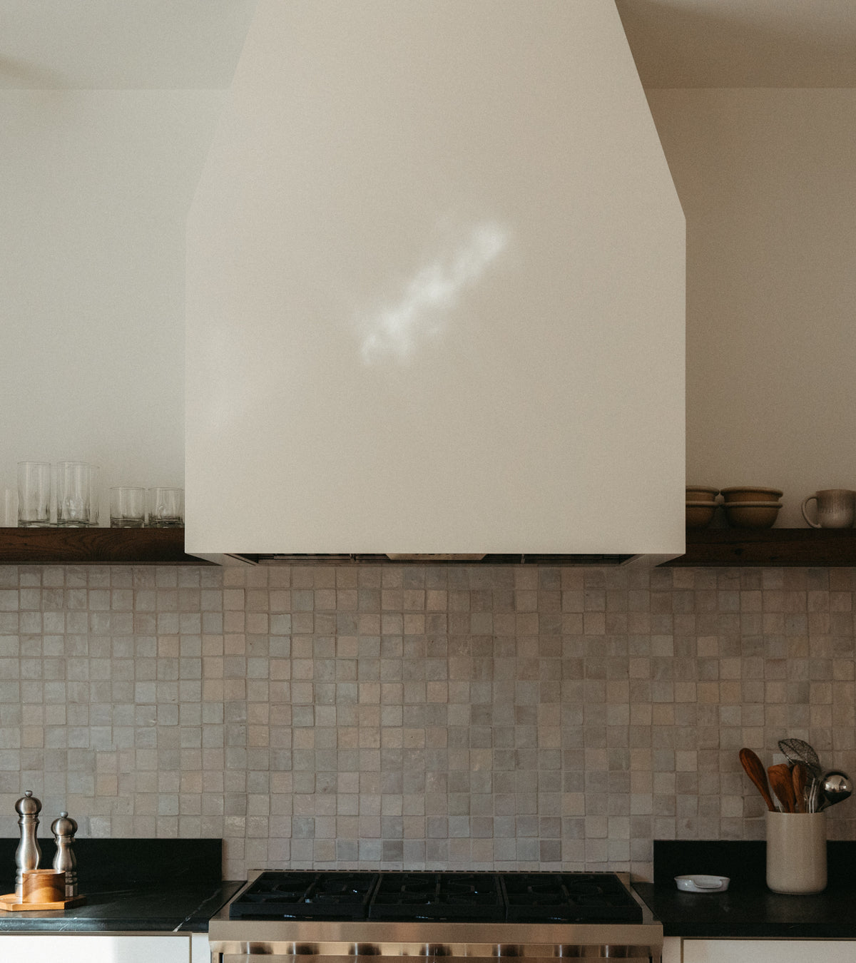 Modern kitchen featuring a Casablanca 2x2 zellige tiled backsplash by Zia Tile, black countertops, gas stove, and large white range hood. Wooden shelf above holds glasses and bowls; utensils and condiments are neatly arranged.