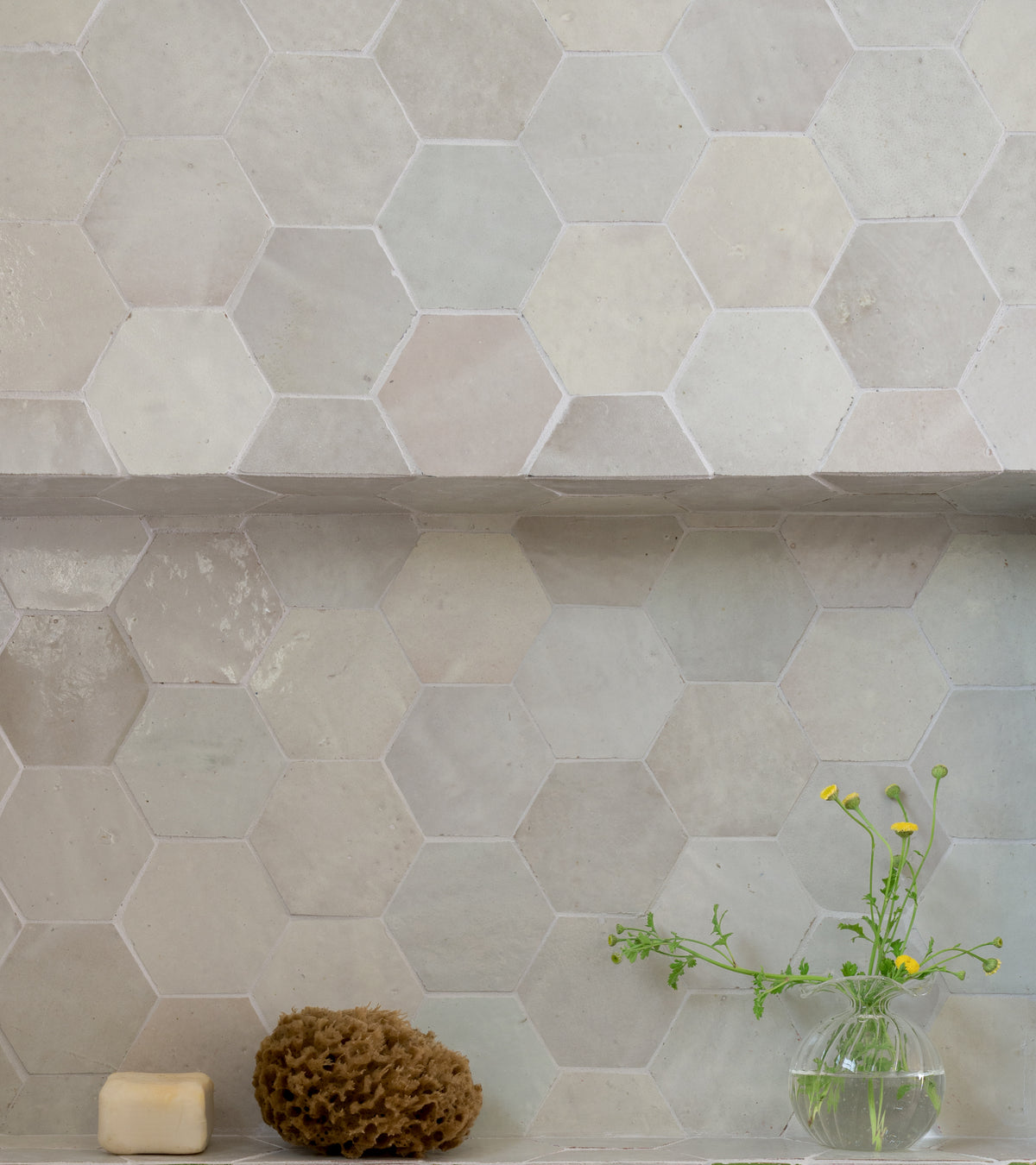 A wall of Casablanca Hex tiles by Zia Tile in light beige features a ledge topped with a natural sponge, bar soap, and a small glass vase holding green stems and yellow flowers.