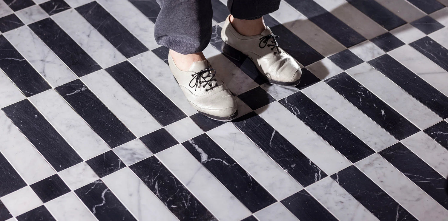 A person wearing white shoes and dark pants stands on a black-and-white striped marble floor, casting shadows. The geometric pattern creates a visually striking effect.