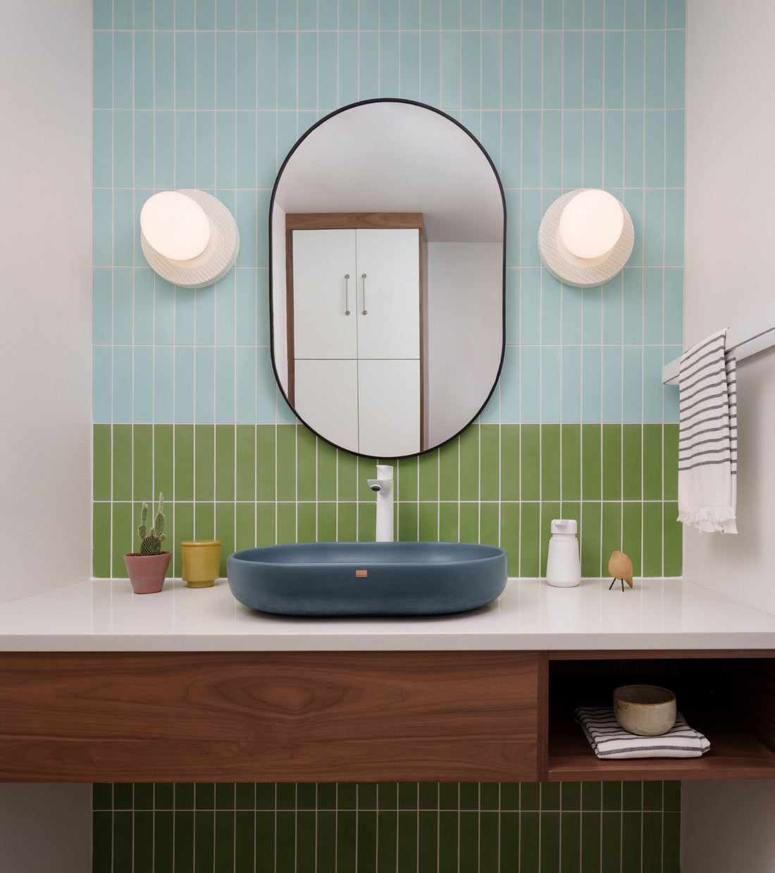 Modern bathroom featuring a blue sink on a wooden vanity, round mirror, two wall sconces, and a Celadon 2x8 subway tile backsplash by vendor-unknown. Plants and soap dispensers bring charm to the space.