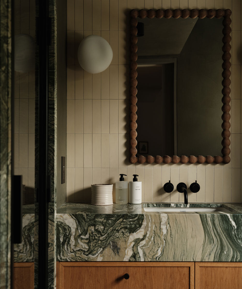A bathroom vanity with a green and white marble countertop, light wood cabinets, two soap dispensers, a textured mirror with a round frame, and beige vertical tile walls. A round wall sconce is mounted beside the mirror.