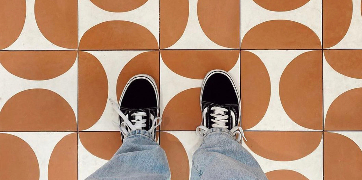 A person wearing black-and-white sneakers and light blue jeans stands on a tiled floor with a geometric orange and white circular pattern.