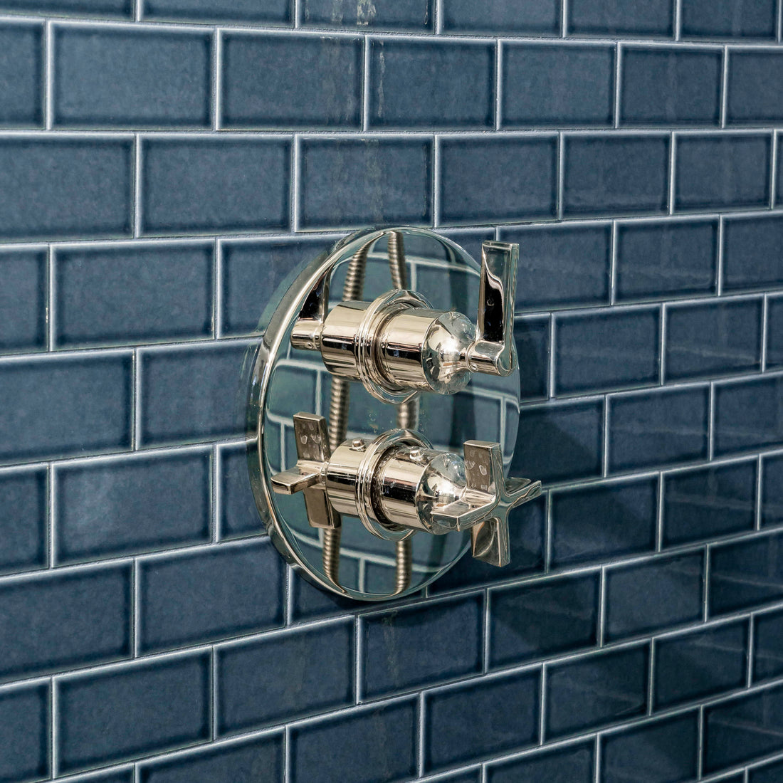 A close-up of a modern, chrome shower control with two handles mounted on a wall covered in glossy, dark blue rectangular tiles arranged in a brick pattern.