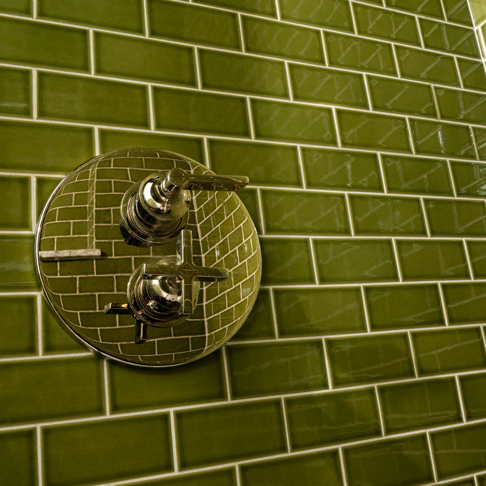 A chrome shower control with two handles is mounted on a wall covered in glossy olive green rectangular tiles, arranged in a brick pattern. The shiny metal reflects the surrounding green tiles.