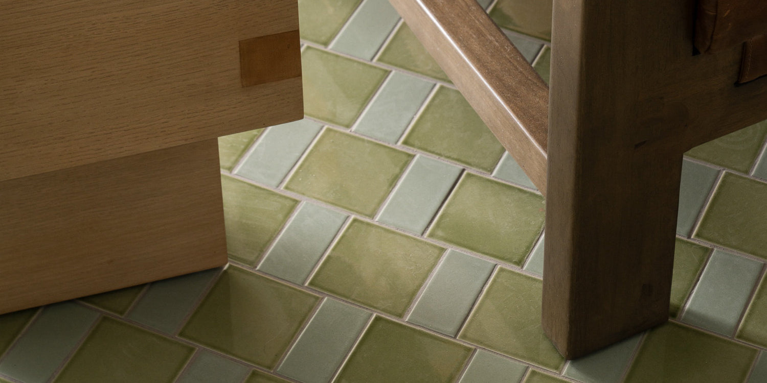 Close-up of green rectangular tiles arranged in a diagonal pattern on the floor, with parts of light wood furniture and a chair leg visible.