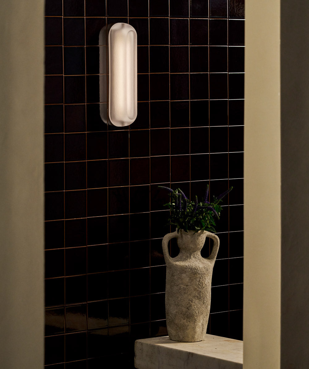 A textured ceramic vase with handles holds small purple flowers on a stone ledge in front of a dark, glossy tiled wall. Above, a modern wall sconce emits a soft, diffused light.