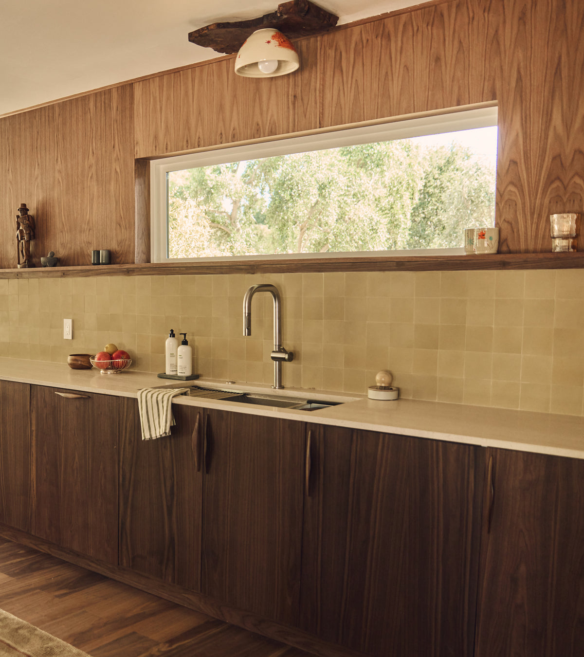 A modern kitchen with wood cabinets features a Clay 4x4 cement tile backsplash from vendor-unknown, beige tiles, a large window above the sink, and sunlight illuminating soap, fruit, and cups on the countertop.