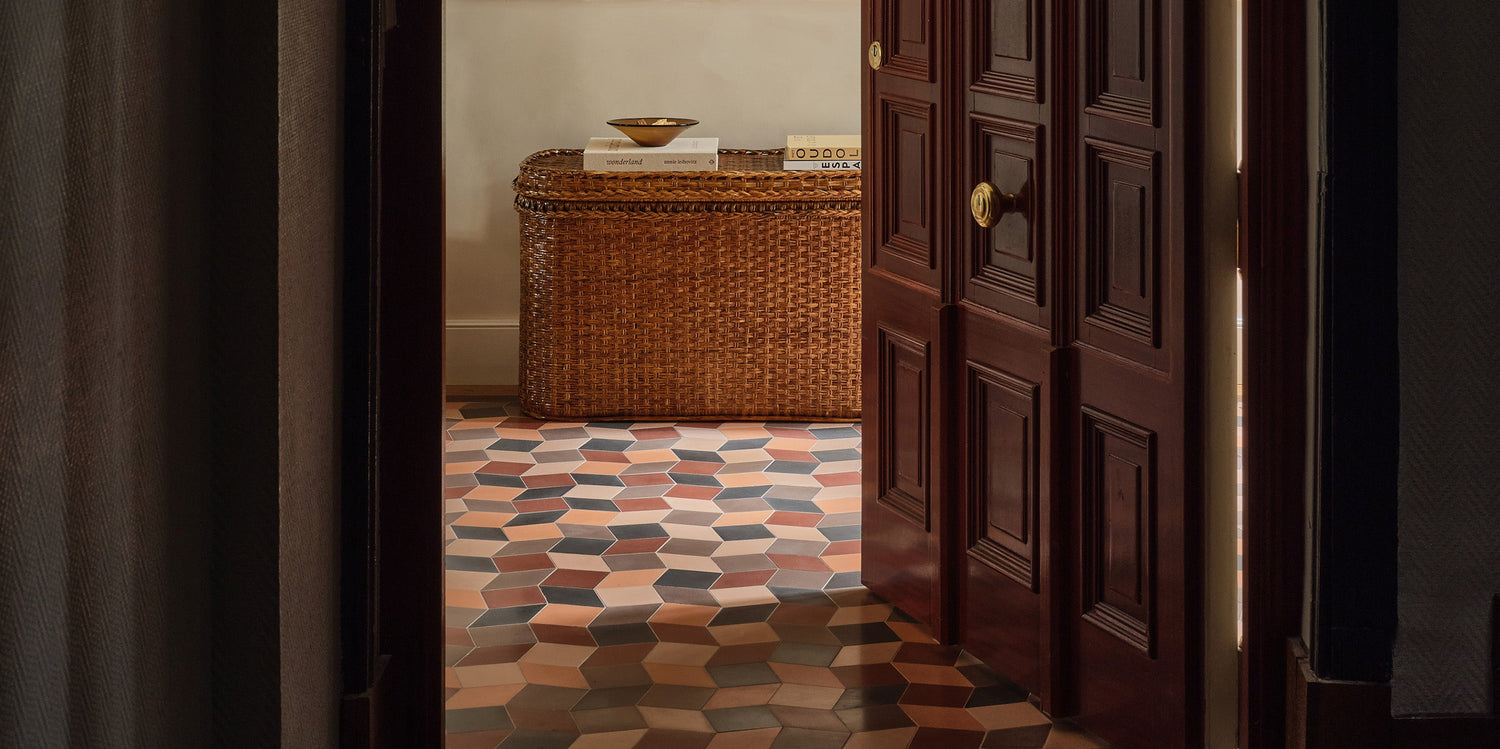 A dark wooden door opens to reveal a room with geometric, multicolored hexagonal floor tiles and a woven wicker chest against a beige wall, topped with a bowl and a few small objects.
