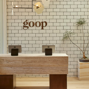 Minimalist reception area with a stone desk crafted from Cotto Allende, two touchscreen registers, a potted plant, and a white tiled wall featuring the goop logo in Zia Tile's Puebla 4x8 Rectangle. Modern hanging light fixtures illuminate the desk.