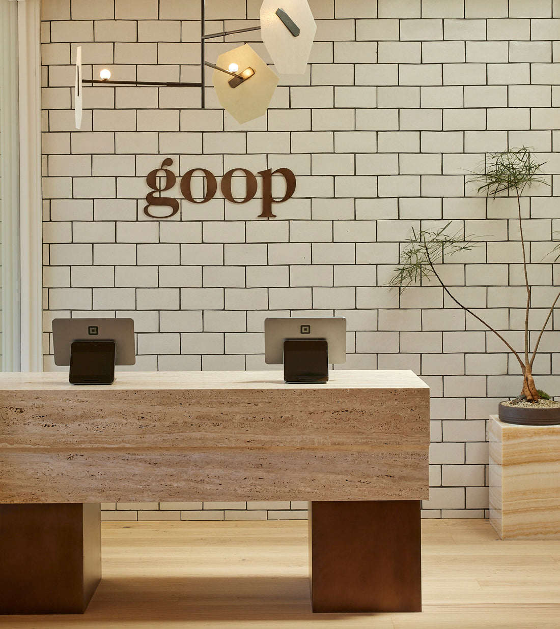 Minimalist reception area with a stone desk crafted from Cotto Allende, two touchscreen registers, a potted plant, and a white tiled wall featuring the goop logo in Zia Tile's Puebla 4x8 Rectangle. Modern hanging light fixtures illuminate the desk.