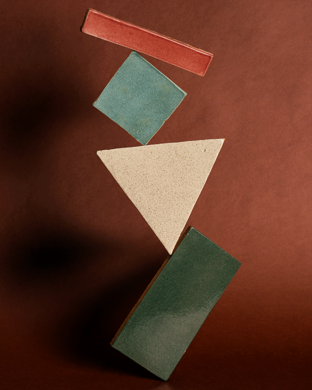 Four geometric tiles—a red rectangle, a blue square, a beige triangle, and a green rectangle—are stacked vertically and appear to balance precariously against a brown background.