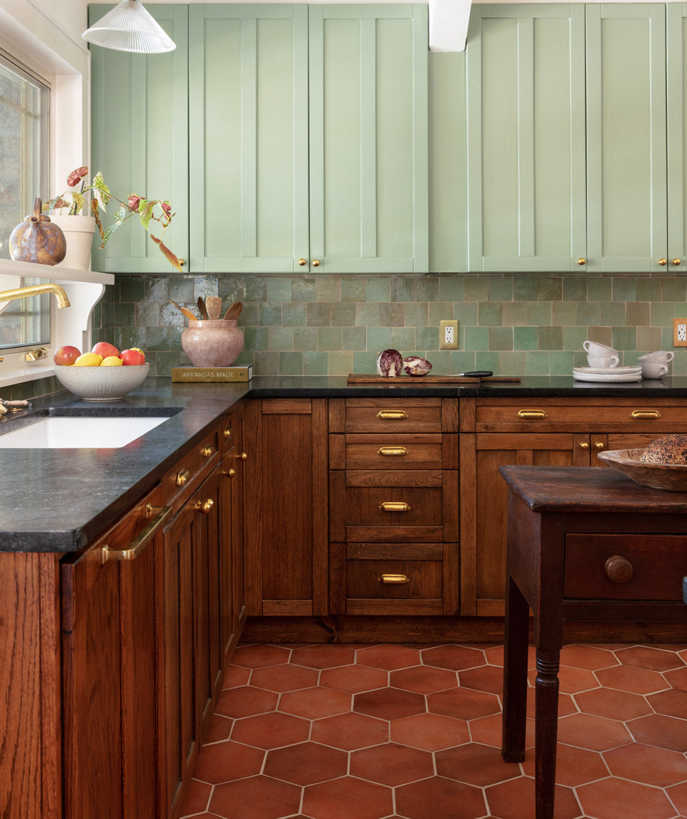 A kitchen with sage green upper cabinets, wooden lower cabinets, brass hardware, green tile backsplash, dark countertops, terracotta hexagon floor tiles, and a window letting in natural light.