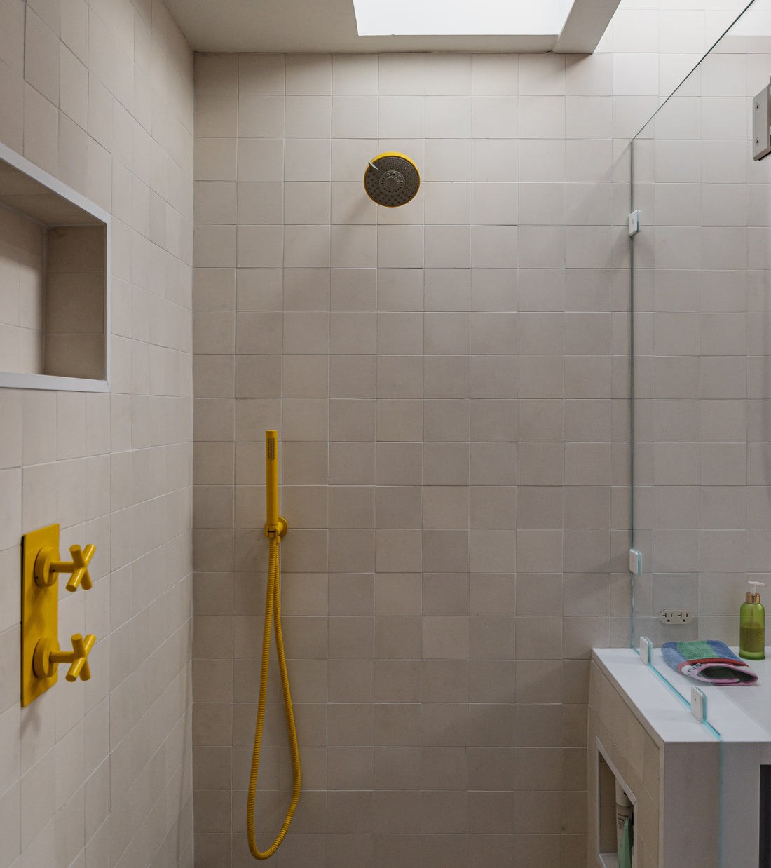 Minimalist shower featuring Cotton 4x4 cement tiles by vendor-unknown in light beige, a yellow showerhead, matching faucet handles, and handheld shower. Glass partition divides the space from a white sink with toiletries; natural light above.