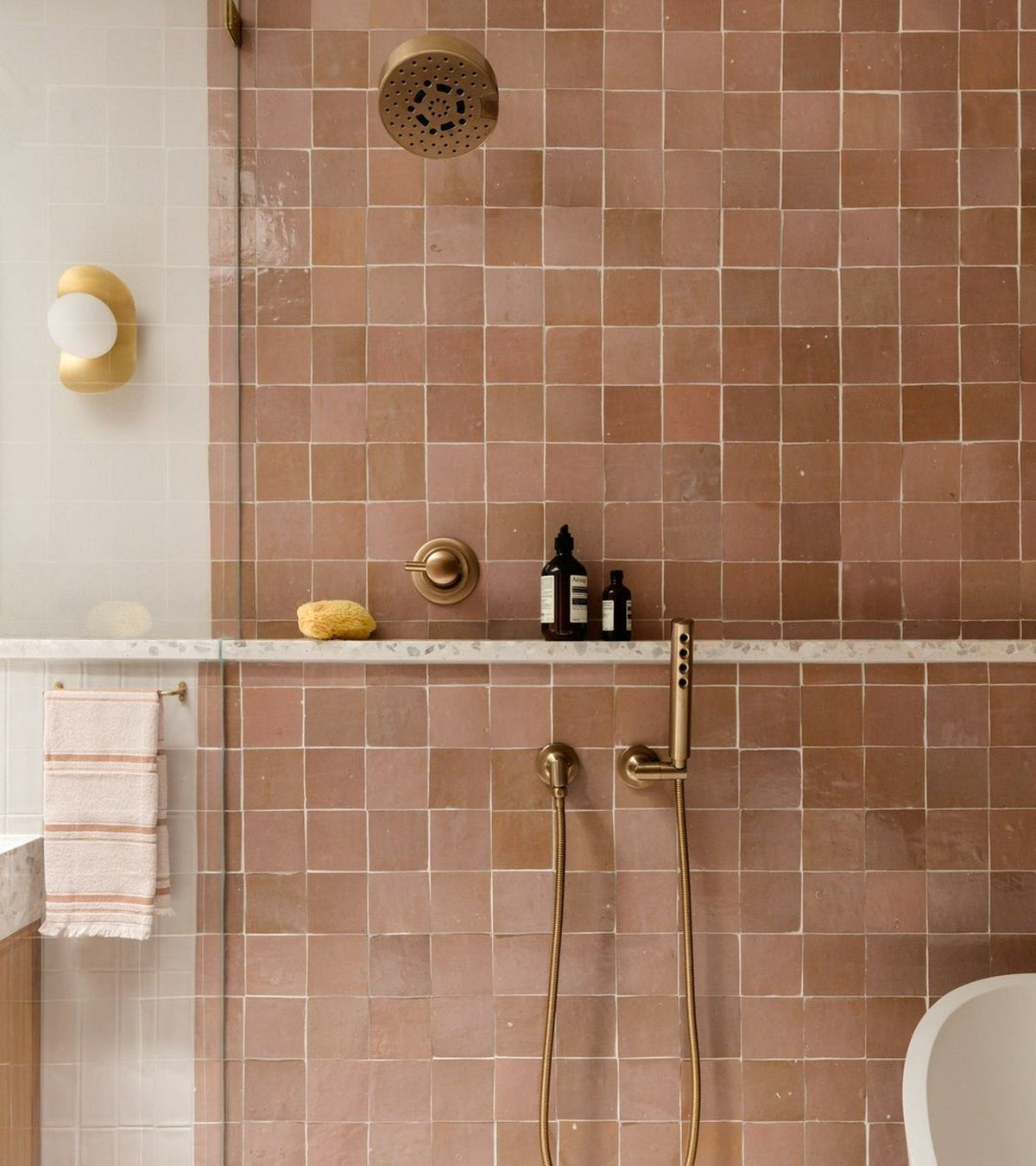 A shower featuring Desert Grey 4x4 tile by vendor-unknown, gold fixtures, a marble shelf with a sponge and bottles, a striped towel hanging, with glimpses of the glass shower door and toilet.