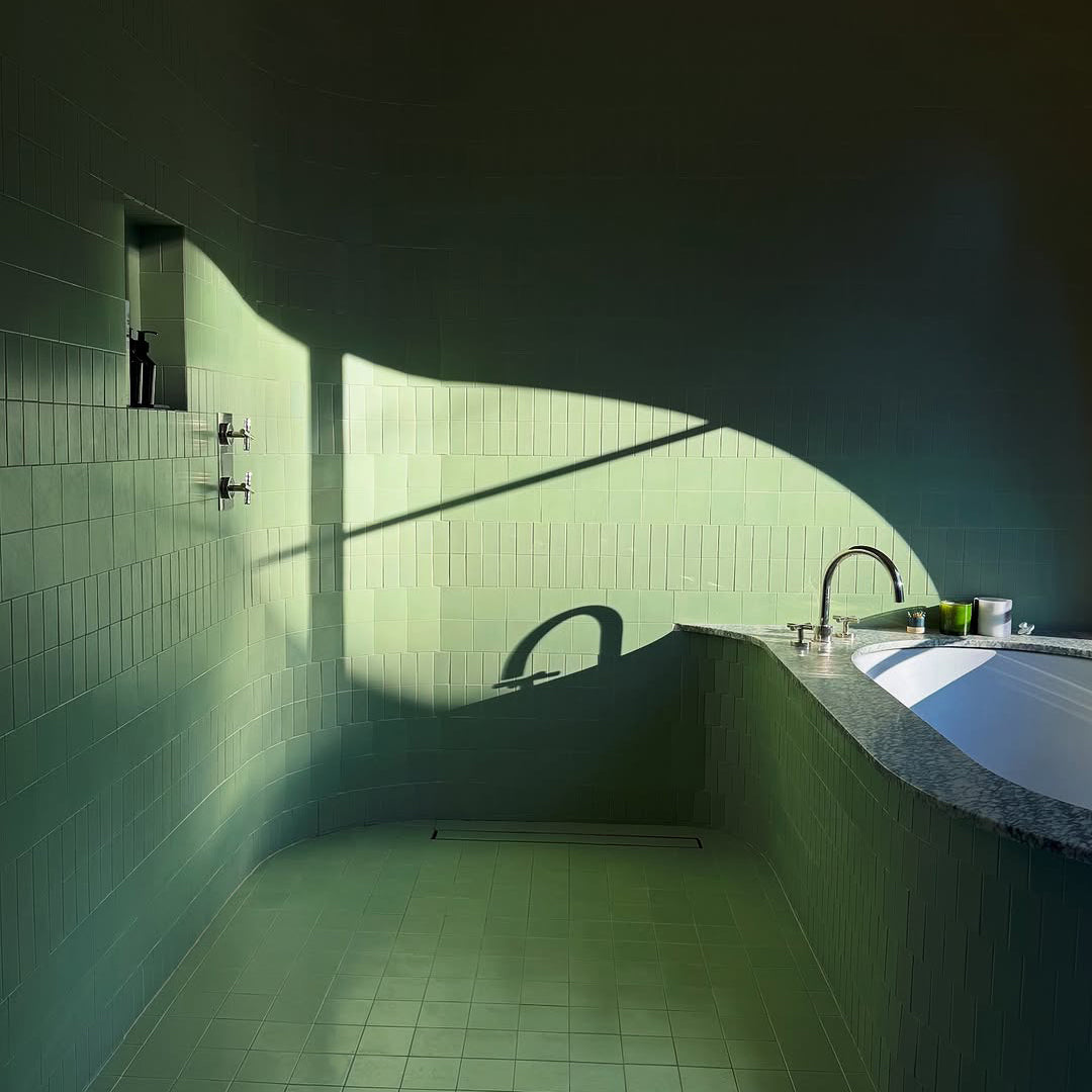 A minimalist bathroom with light green tiled walls and floor, a sunken white bathtub with a granite edge, and dramatic sunlight casting shadows across the curved wall and floor.