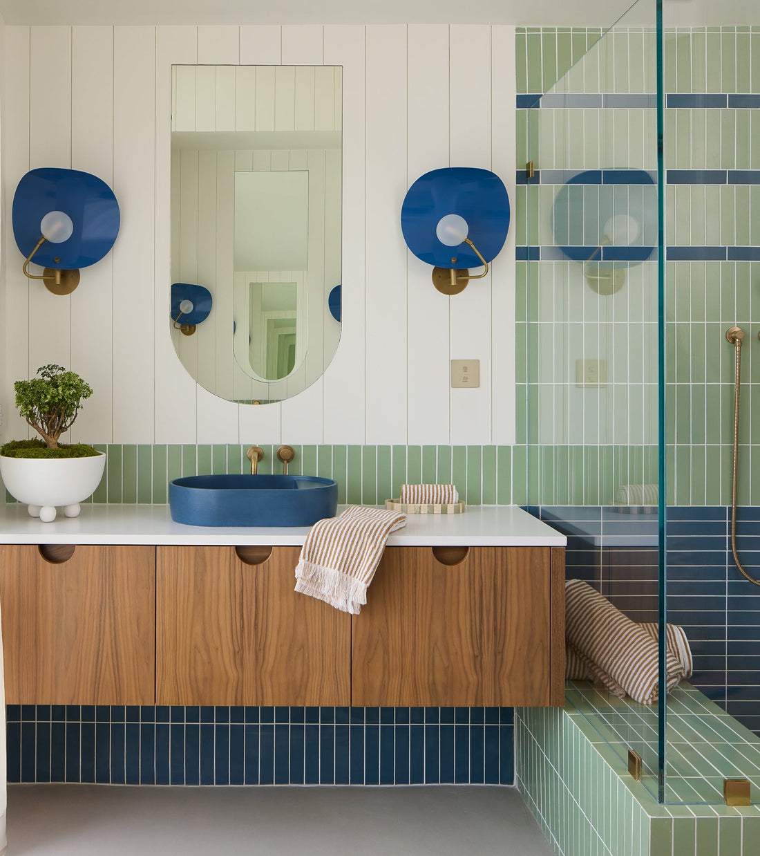 Modern bathroom with wood vanity, oval mirror, blue circular sink, striped towels, and blue sconces. Zia Tile's Eucalyptus 2x8 ceramics in green-blue cover the shower and wall; a small potted plant sits on the counter.