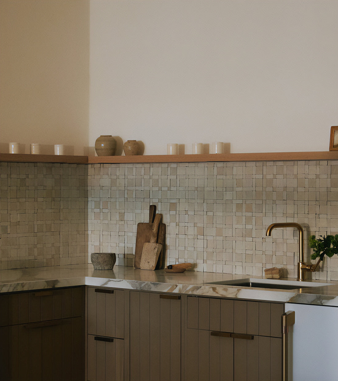 A modern kitchen featuring brown cabinets, a marble countertop, gold faucet, and Gambit 1 mosaic zellige tile backsplash by Zia Tile. Wooden cutting boards, jars, and candles decorate the counter and open shelf.