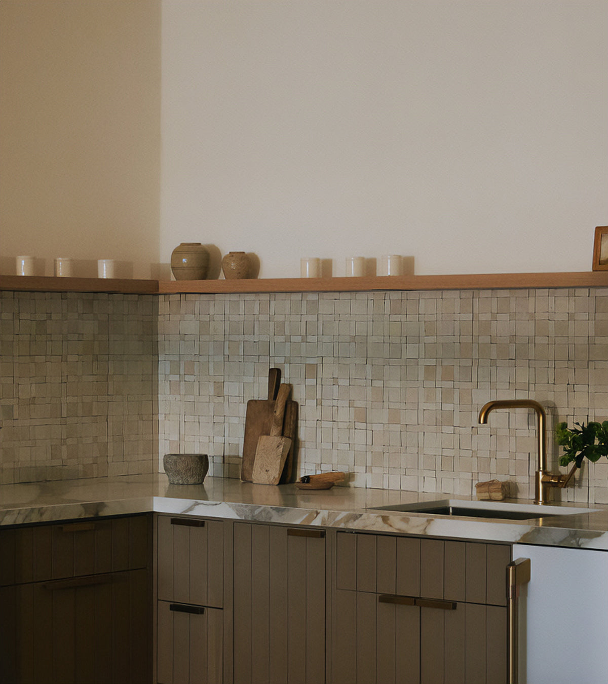 A modern kitchen featuring brown cabinets, a marble countertop, gold faucet, and Gambit 1 mosaic zellige tile backsplash by Zia Tile. Wooden cutting boards, jars, and candles decorate the counter and open shelf.