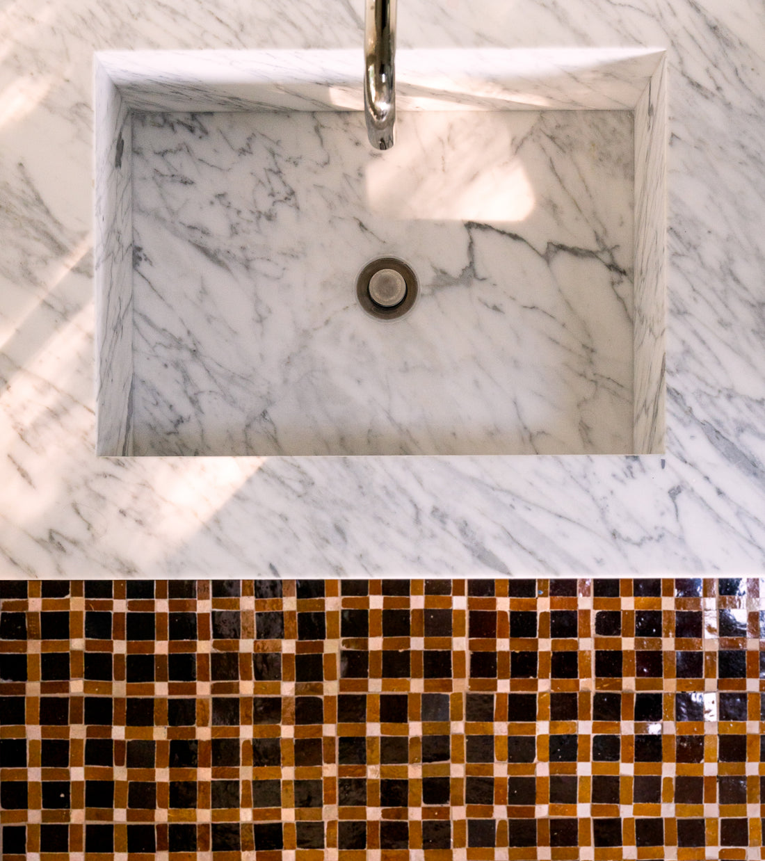A top view of a rectangular marble sink with silver faucet sits on a white countertop with grey veining. The lower edge features Zia Tile’s Gambit 5 mosaic zellige tiles in alternating brown, black, and gold as the backsplash.