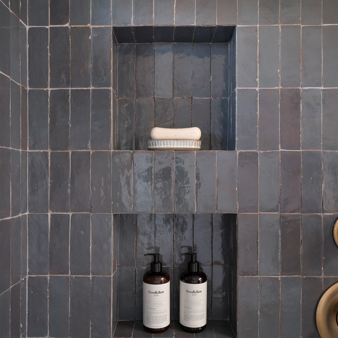 A shower niche with dark blue tiles holds a white soap dish on the top shelf and two dark pump bottles with white labels on the bottom shelf. A brass shower control is partially visible on the right.